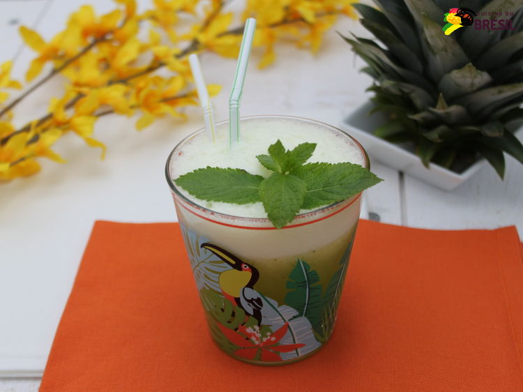 Un verre de jus d'ananas et menthe sur un fond orange entouré d'un bouquet de fleurs jaune et d'une déco des feuilles d'ananas